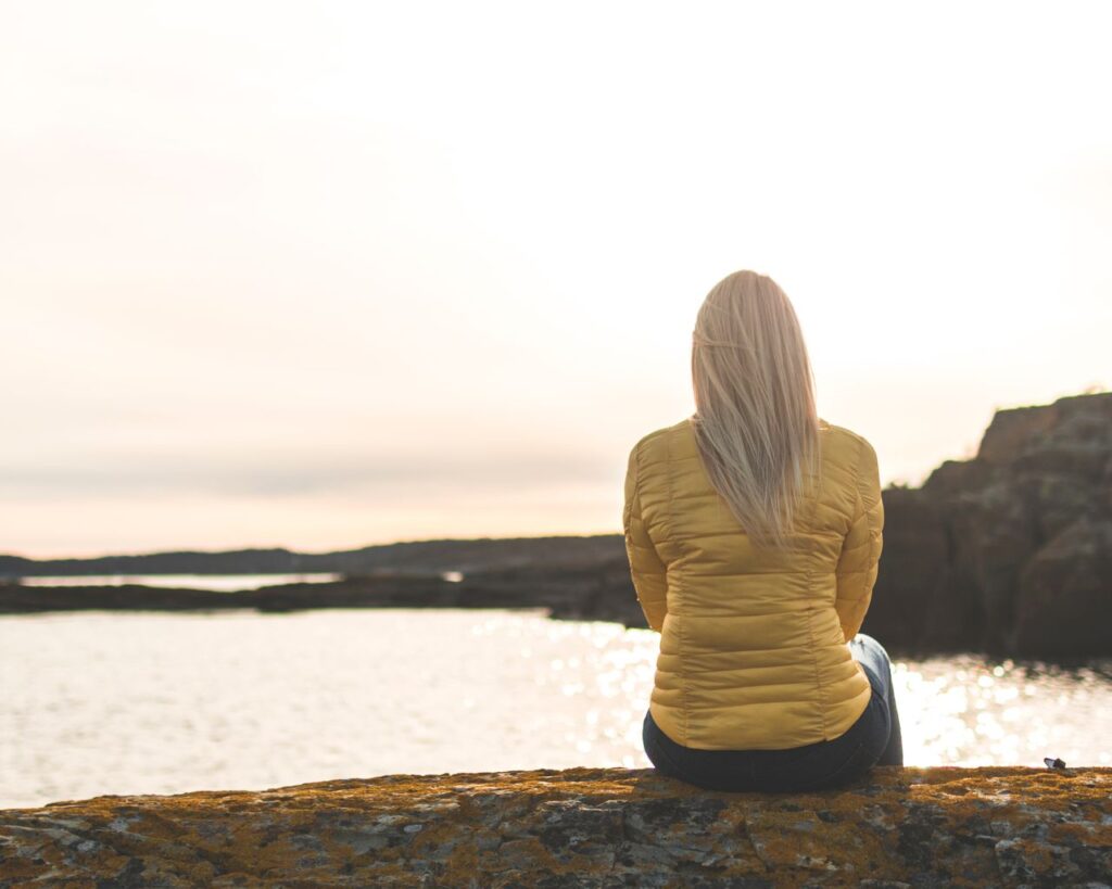 A peaceful scene of a woman in a yellow jacket sitting by the ocean at sunset, reflecting in solitude.
