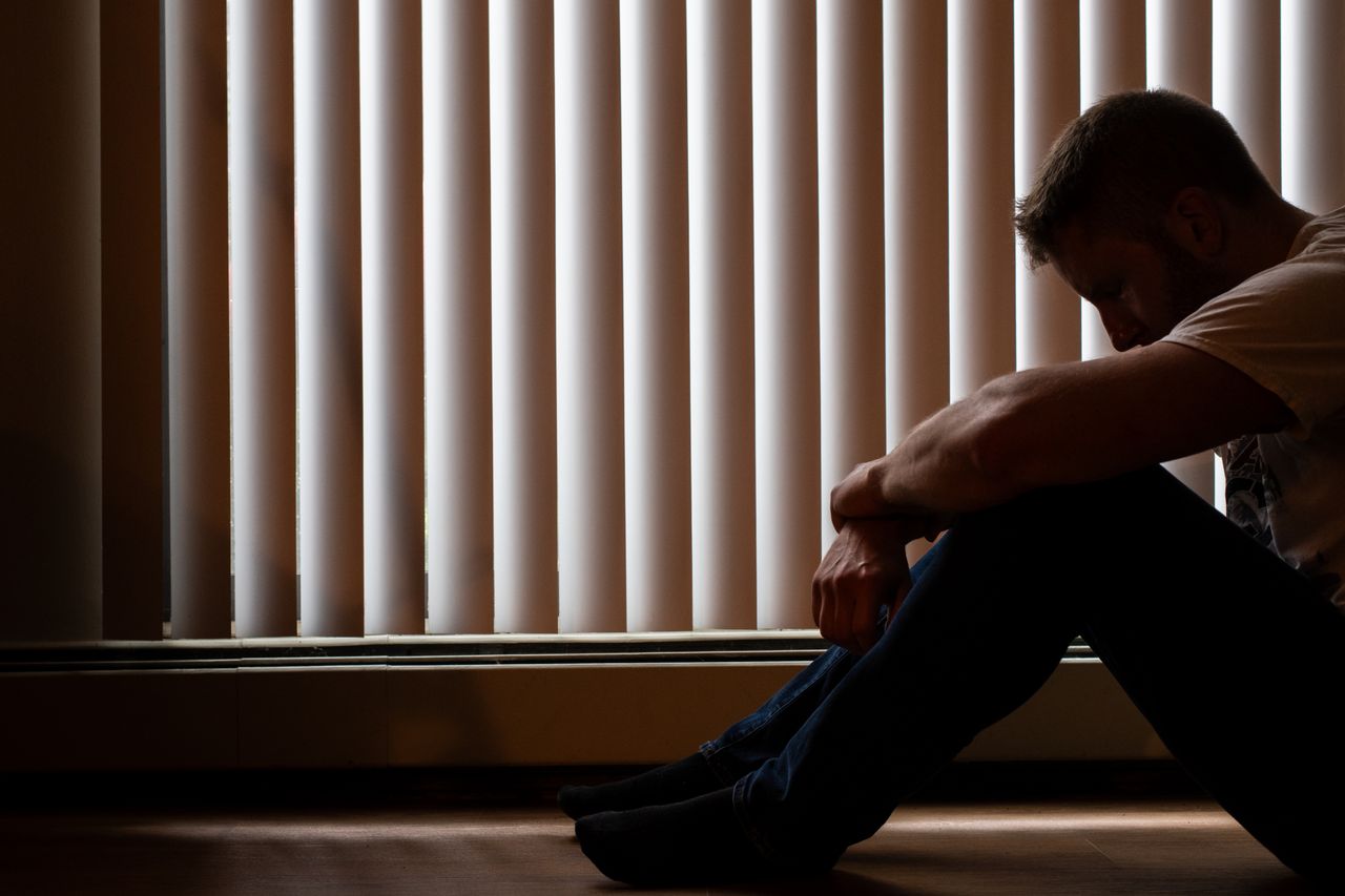 A man sits silhouetted against window blinds, conveying solitude and introspection.