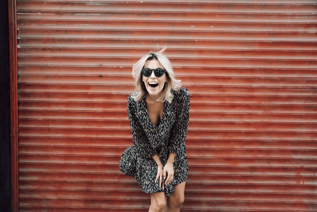 Positive female wearing modern sunglasses and dress standing near shutter and laughing happily